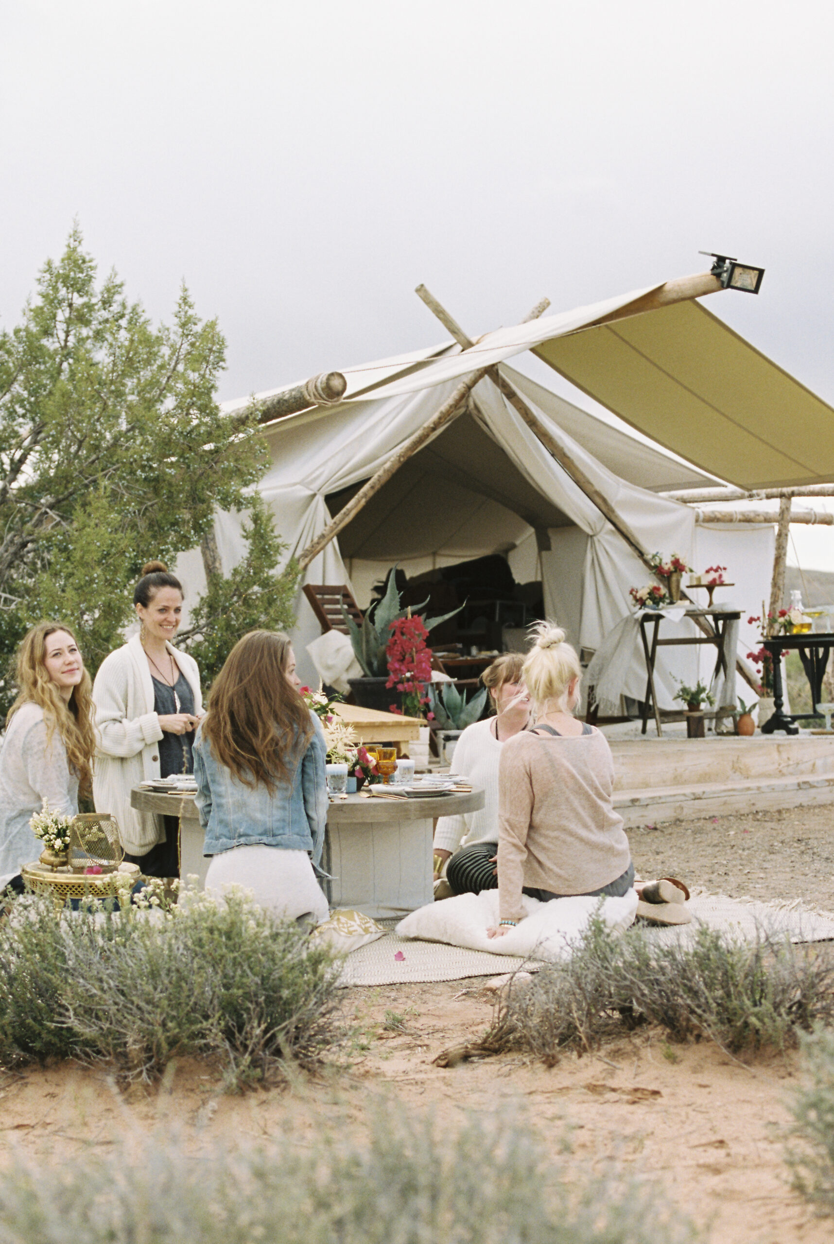 Group of women friends sitting on the ground round a table outside a tent in the background.